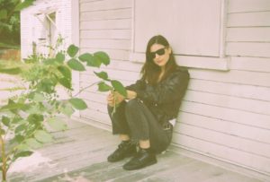 Woman wearing all black and sunglasses leaning against white house.