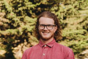 Man with red curly hair and glasses in red shirt in front of trees.