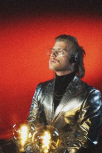 Man in silver jacket with glasses and headphones on staring off to right.