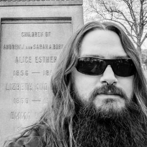 Man with long hair, beard, wearing glasses standing in front of a tombstone.