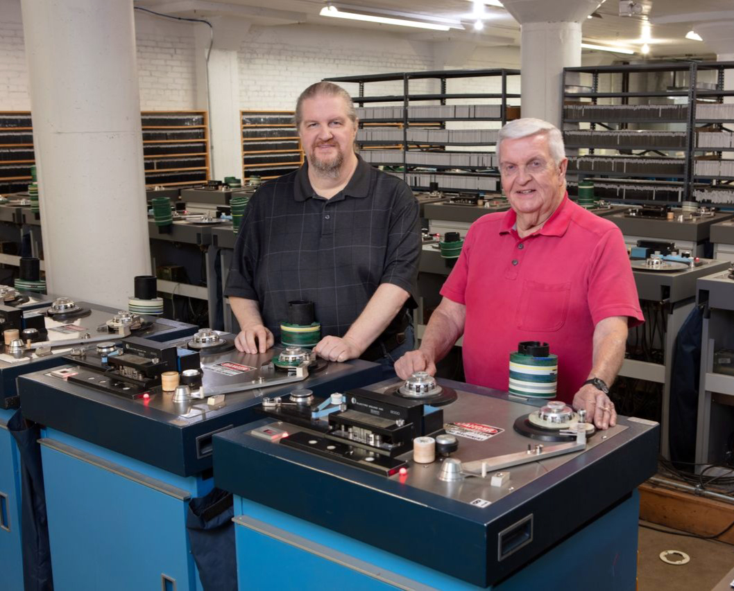 two men standing next to a tape duplication machine